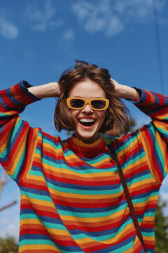 Woman, smiling, striped sweater, sunglasses, short brown hair, outdoor, blue sky, joyful, 20s model in colorful knit pullover laughing with hands in hair, casual fashion lifestyle travel vibe