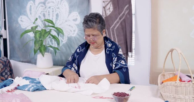 Senior Asian woman artisan works in home studio, threading needle and stitching resist pattern on cloth for shibori indigo dye, sustainable craftsmanship scene for heritage craft revival story.