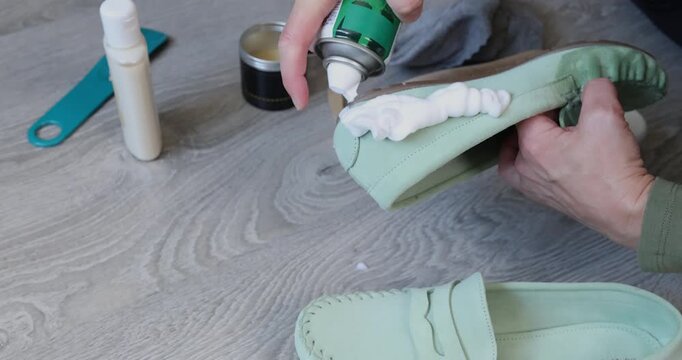Person applying white cleaning foam from spray can on light green leather loafers. Process of footwear care with professional cleaner on grey wooden background. Hands cleaning elegant suede shoes