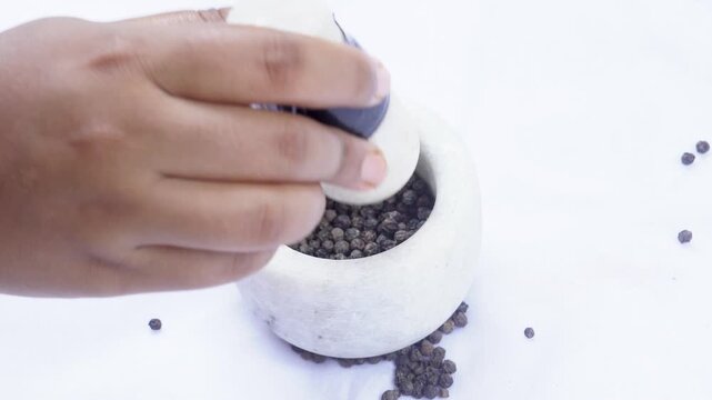 Grinding black whole pepper in a white stone mortar. The hand holds a pestle and pounds black pepper in a circular stone mortar isolated on a white background.