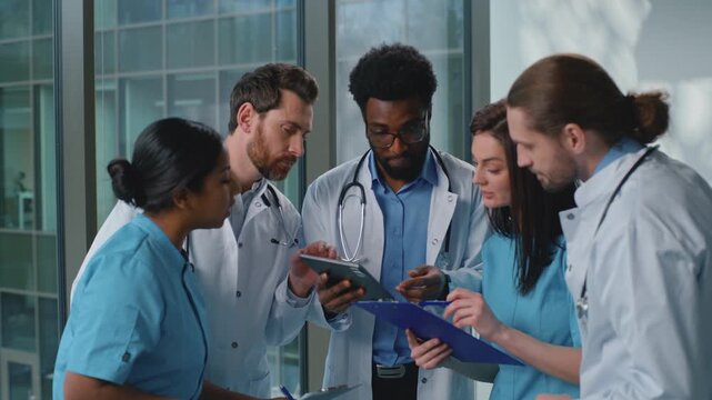 Focused multiethnic five doctors with digital tablet working talking deciding medicine diagnosis in clinic hallway. Multiracial nurses practitioners therapists surgeons talk team work cooperative