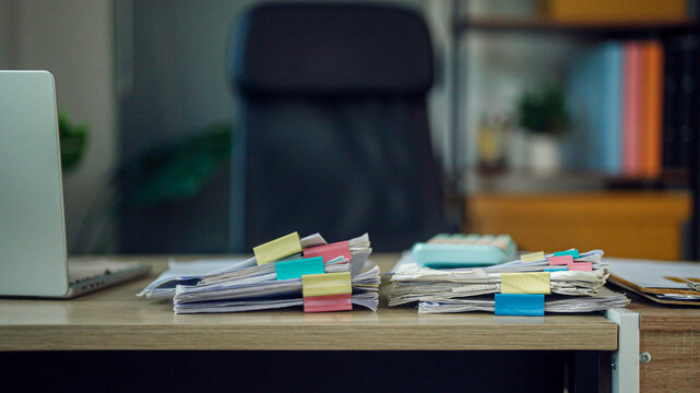 Cluttered desk holds stacks of papers and colorful sticky notes. Laptop and chair suggest a busy, active workspace. Office environment shows organization challenges