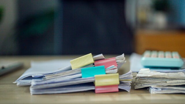 Stacks of papers hold colorful clips. Calculator rests nearby, hinting at finance. Blurred background suggests office setting. Focus sharpens on documents and their labels