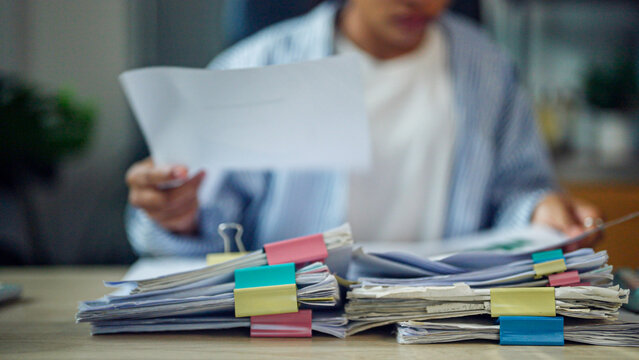 Person reviews documents with focused expression. Stacks of papers marked with colorful clips. Desk clutter shows heavy workload. Professional setting implies deadline pressure