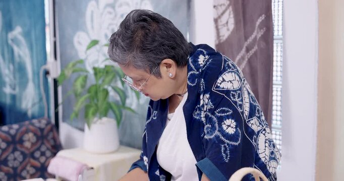 Senior Asian woman artisan bends close, reaches toward worktable supplies, continuing resist stitching preparation on cotton cloth for shibori indigo dye, home studio family small business workflow.