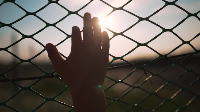 Hand touching chainlink fence at sunset with fingers on wire mesh. Person places hand on fence barrier outdoors. Silhouette shows hand at chainlink. Fingers touching wire fence at sunset view.
