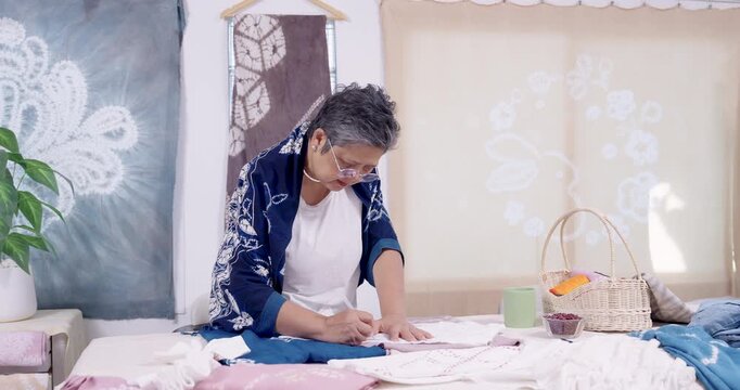 Senior Asian artisan writes pattern marks on cloth, measuring layout for resist stitching, preparing shibori tie dye textile in home studio heritage craft revival for sustainable small business brand.