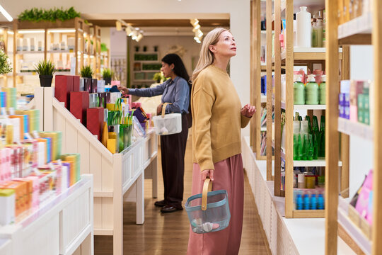 Caucasian middle aged woman browsing cosmetic products in beauty store, comparing skincare options and retail choices. Useful for beauty retail, consumer shopping, family purchase