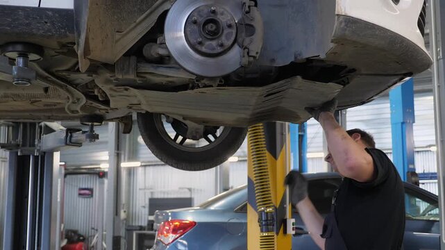 A professional car mechanic repairs a car on a lift. He uses a screwdriver and a hammer to do so. This is how modern auto repair shops work.
