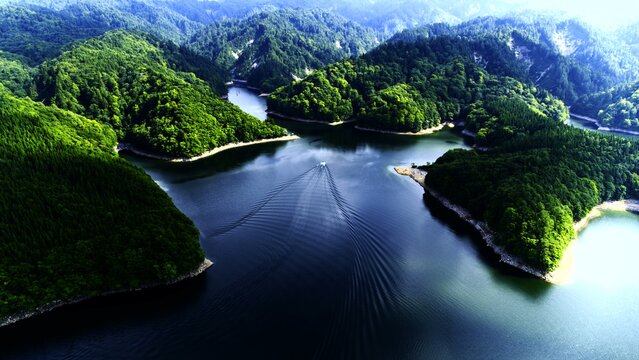 This expansive landscape shows a serpentine lake winding through lush green forested hills with a small boat traveling on the water leaving a trail under bright daylight in a serene environment