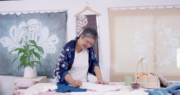 Senior Asian woman artisan leans over table, marks shibori pattern on fabric with pen, preparing resist stitching guide before indigo dyeing, sustainable handmade textile for boutique customer order.