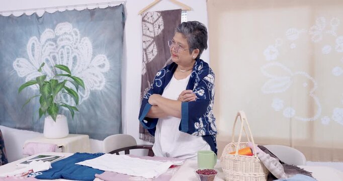 Senior Asian retiree craft entrepreneur stands beside worktable, folds arm, studies shibori fabric pieces, compares pattern option, prepares indigo dyeing plan for customer order in home studio.