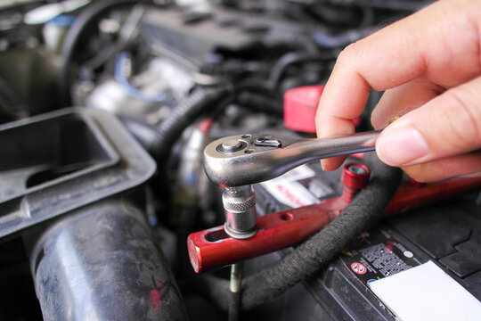 Hand using a socket ratchet wrench to tighten a bolt on a red car battery tie-down bracket during automotive engine maintenance