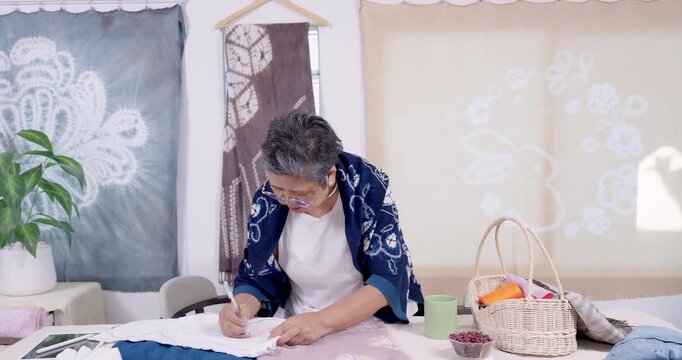 Senior Asian artisan folds finished fabric bundle, stacks shibori panel for indigo tie dye batch, checks cotton texture, prepares inventory for slow fashion brand in small business studio today.