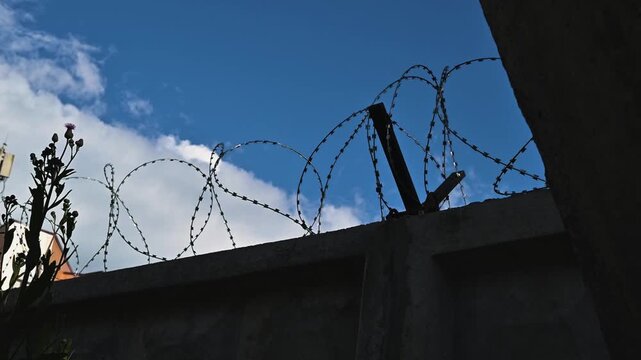 Barbed wire is installed at the top of a wall under a blue sky with clouds. The setting suggests a place that requires protection and security