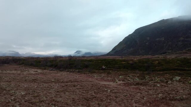 Tracking extreme wide shot of a woman riding a horse along a trail in a barren valley, with mist-covered mountains and a cloudy sky surrounding the scene for a cinematic wilderness view.