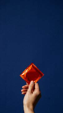 A male hand holds a bright orange condom wrapper against a solid dark blue studio background highlighting safe sex awareness and reproductive health care options
