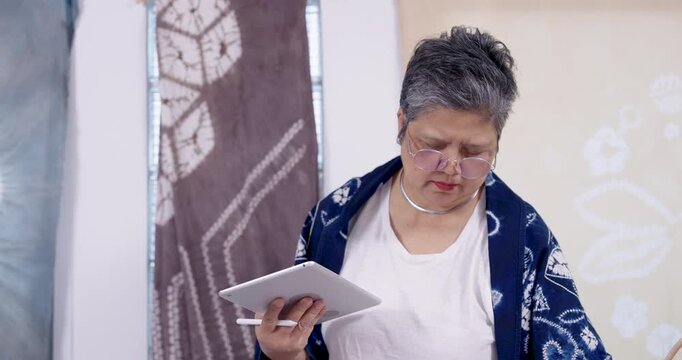 Senior Asian woman artisan studies tablet screen in close view, reviews pattern sketch, confirms measurements, prepares cotton cloth for shibori resist stitching, steady slow craft planning moment.