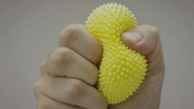 Hand squeezing a textured yellow stress ball in a neutral background, showcasing the gradual compression and deformation of the object in three sequential frames