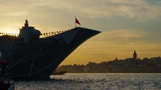 A massive grey naval vessel bow dominates the foreground in Istanbul, set against a golden sunset skyline and the historic cityscape of the Bosphorus strait