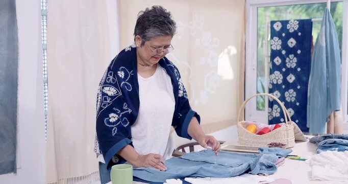 Senior Asian woman artisan walks around worktable, checks denim panel layout, plans shibori resist stitching sequence, prepares cotton fabric for indigo dyeing, home studio workshop workflow.