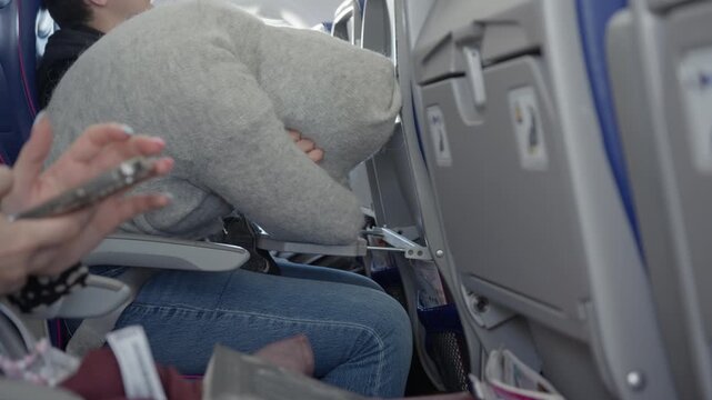 Tired passenger sleeps with folded arms on an airplane cabin tray table during a long transit flight while another person sits in an economy class seat using a wireless mobile smartphone.