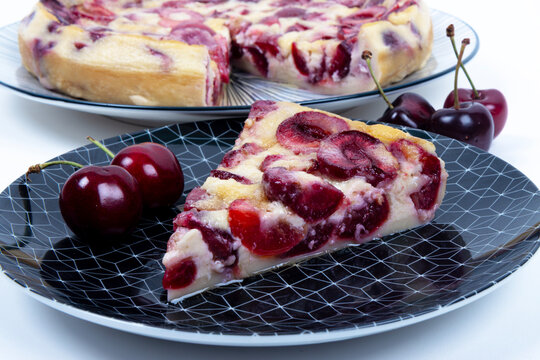 Part de clafoutis aux cerises fait maison sur une assiette noire &agrave; motifs avec des cerises fra&icirc;ches