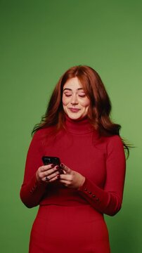 A young woman in a red dress smiles while looking at her phone against a green screen background.