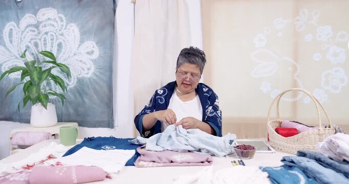 Wide view shows senior Asian woman artisan at table, stitches denim with red thread, builds shibori resist pattern, tool and fabric arranged, workshop routine for sustainable family small business.