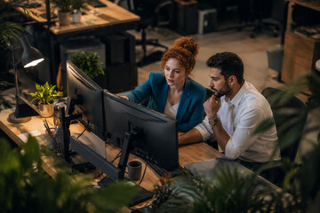 Fototapeta premium Two colleagues working late at modern office, focused on computer screens, surrounded by plants