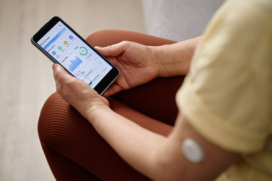 Close up of woman holding smartphone and checking glucose tracking app with arm sensor, managing diabetes care. Ideal for telehealth, mobile health, chronic care marketing