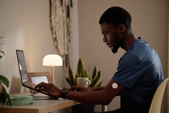 Black young adult man working on laptop at home, monitoring glucose sensor on arm and managing digital health data. Side view, ideal for diabetes care, telehealth, wellness campaigns