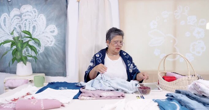 Senior Asian woman artisan continues needlework on denim, secures resist stitch, checks pattern progress, prepares next section for indigo dyeing, quiet studio workflow for handmade textile order.