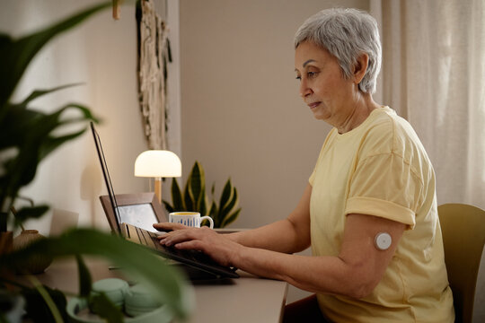 Senior Asian woman using laptop at home, managing online tasks while wearing glucose sensor, representing diabetes monitoring and digital healthcare. Side view for telehealth marketing