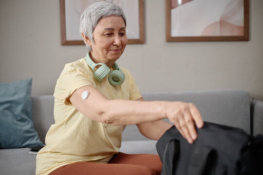 Senior Asian woman packing gym bag at home, showing glucose sensor and preparing for workout, representing active diabetes management. Ideal for wellness, healthcare, fitness campaigns