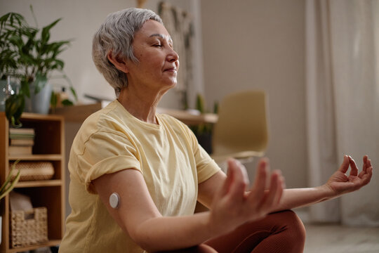 Senior Asian woman meditating with eyes closed at home, wearing glucose sensor and practicing mindful wellness routine. Ideal for diabetes care, healthy aging, self care marketing