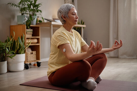 Middle aged Asian woman meditating on yoga mat at home, wearing glucose sensor and practicing mindful wellness routine. Ideal for diabetes management, healthy lifestyle, self care
