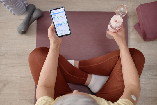 Overhead shot of senior woman checking glucose tracking app while holding water bottle on yoga mat, managing diabetes and fitness routine. Ideal for wellness, telehealth, prevention marketing