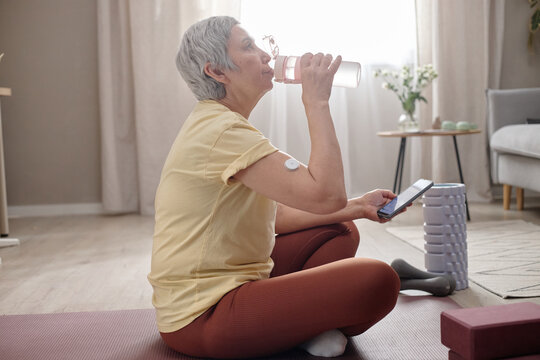 Senior Asian woman resting after home workout, drinking water and checking smartphone with glucose sensor on arm, showing active diabetes management. For wellness, fitness, healthcare ads