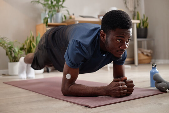 Young Black man holding forearm plank on exercise mat at home, tracking fitness with wearable sensor and building strength. Ideal for wellness, home workout, health tech marketing