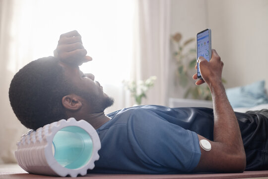 African American young adult man resting after workout, checking fitness app on smartphone with glucose sensor on arm, using recovery routine. Ideal for wellness, diabetes, fitness marketing