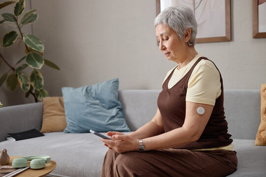 Senior Asian woman checking smartphone while monitoring glucose sensor on arm at home, managing diabetes and digital health routine. Useful for telehealth, chronic care, wellness marketing