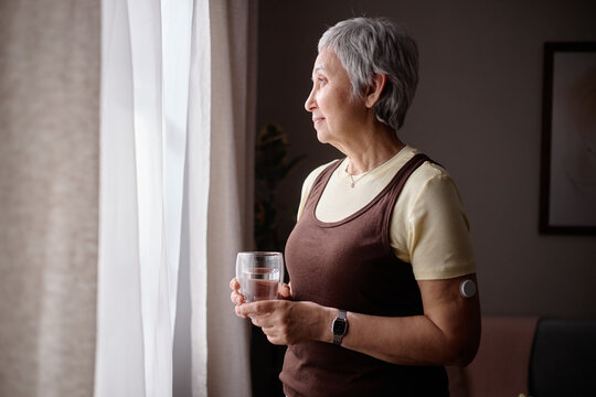 Senior Asian woman holding glass of water and looking through window, showing glucose sensor and wellness monitoring. Useful for diabetes care, healthy aging, home healthcare