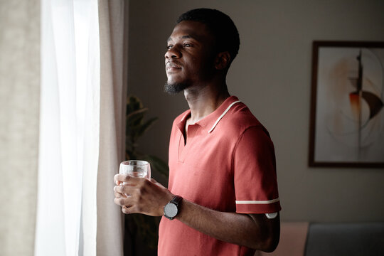 Medium shot of young adult Black man holding glass of water and looking through window, reflecting on daily routine and wellbeing. Ideal for wellness, hydration, mindfulness campaigns