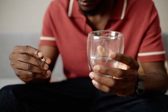Close up of Black young adult man holding pill and glass of water, preparing medication at home, showing healthcare treatment and self care. Useful for pharmacy or wellness campaigns