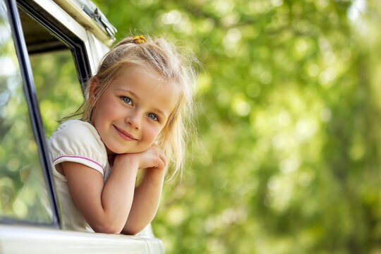 Adorable young blonde little girl with blue eyes and long hair smiling brightly while looking, leaning out of a car window during a sunny summer road trip in the countryside in nature