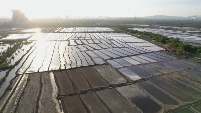 A wide drone aerial movement shot of the sun reflecting in expansive water filled salt pan beds or flats and an urban landscape visible in the background in the evening, Mumbai, India