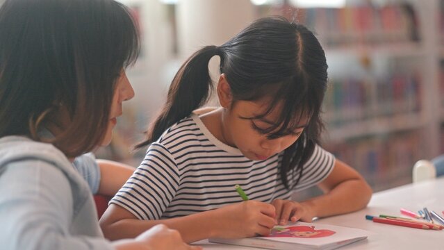 Clever Asian youth learner reading textbook chapters with classmate friend at library table. Clever little girls collaborating on educational school homework to build confidence.
