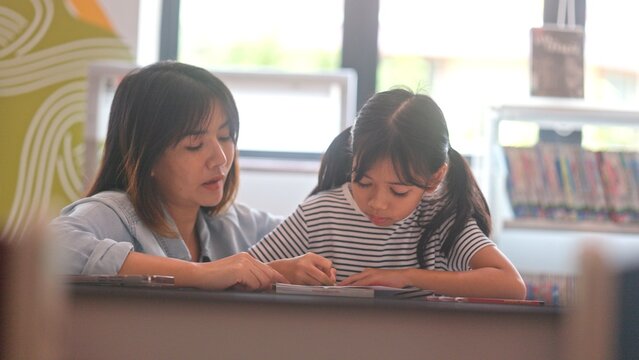Cute Asian girl child discussing book stories with classmate friend at library table. Clever little girls collaborating on educational school homework to enjoy group learning.