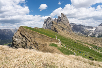 Breathtaking panoramic view of the iconic Seceda mountain ridge with hikers walking along paths on green pastures under a cloudy blue sky, alpine travel concept © Rokas